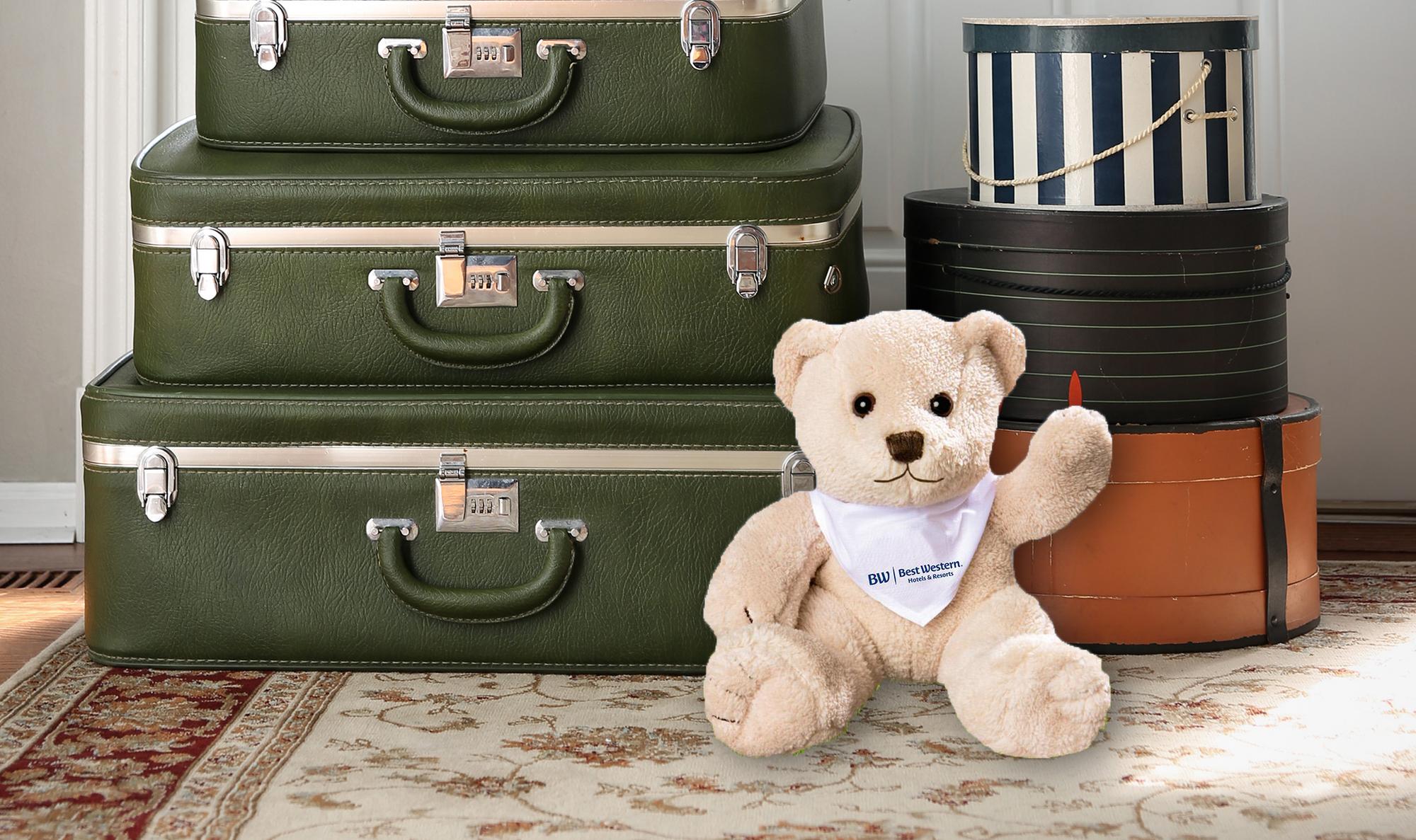Stoffbär sitzt vor gestapelten Koffern und Hutschachteln auf einem Teppich.