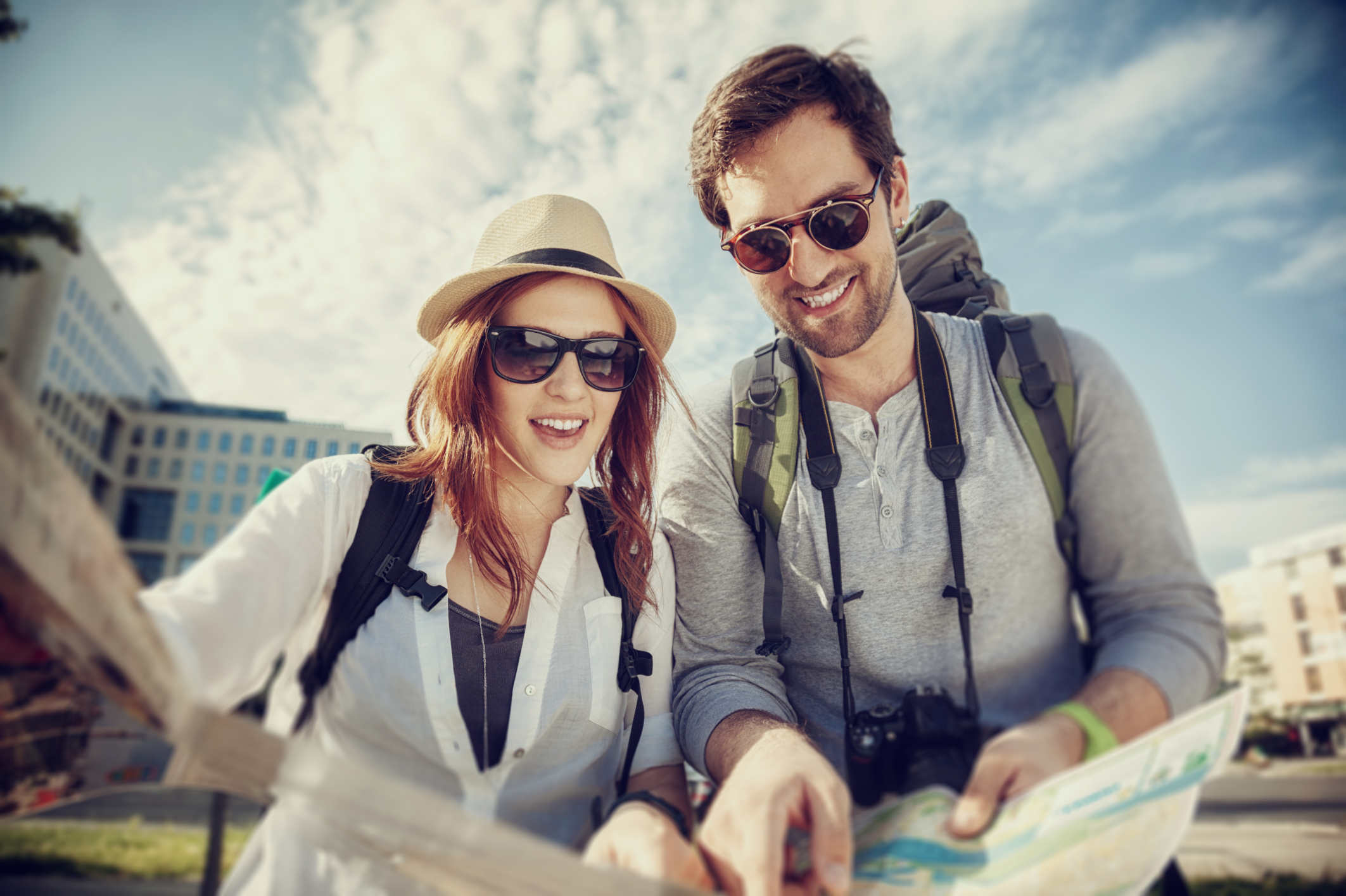 Zwei Reisende mit Rucks&auml;cken und Karte erkunden eine Stadt bei teils bew&ouml;lktem Himmel.