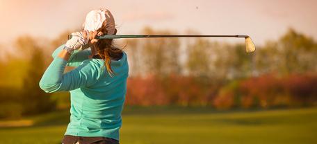 Golfer beim Abschlag auf grünem Platz mit Bäumen im Hintergrund bei Sonnenlicht.