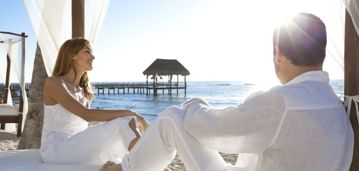 Paar entspannt am Strand in wei&szlig;er Kleidung mit Blick auf Meer und Steg.