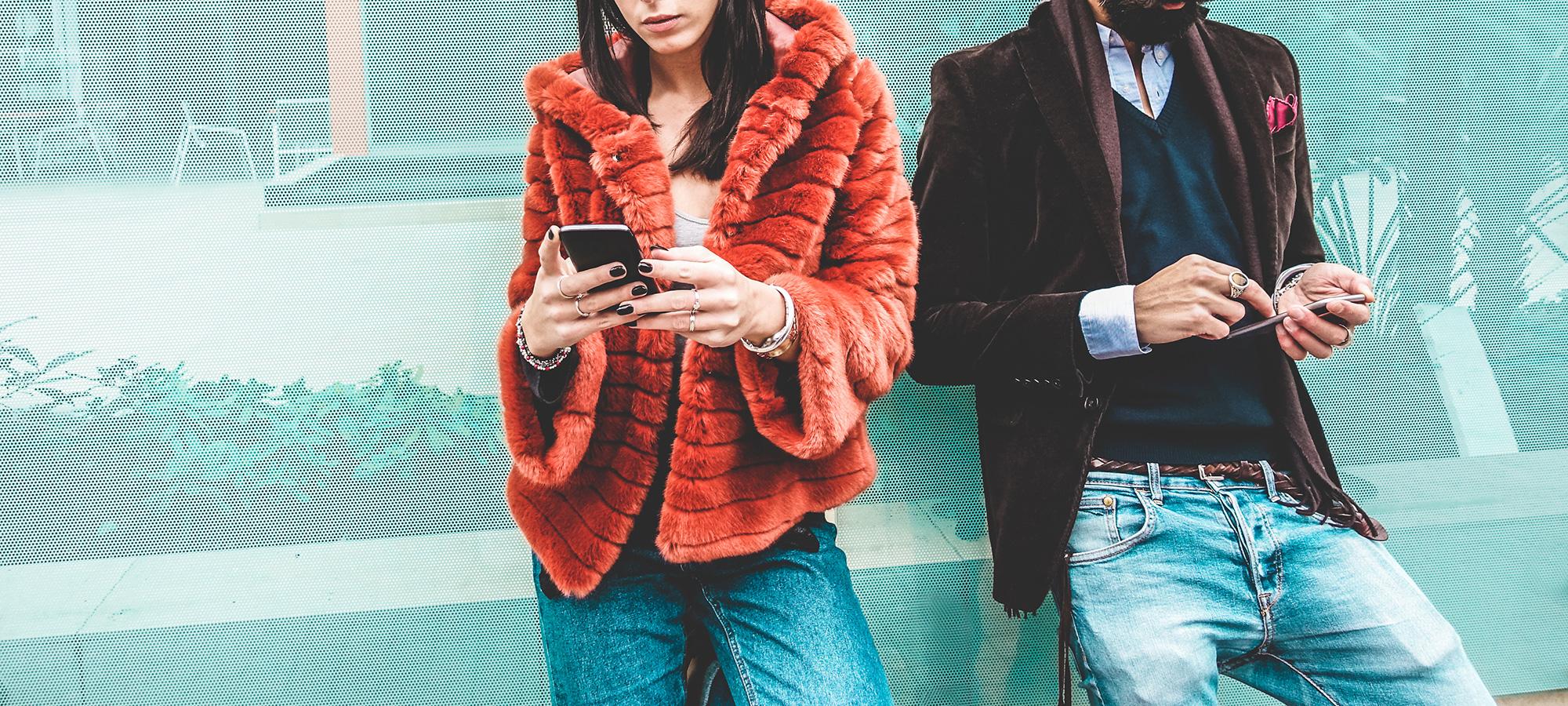 Zwei Personen lehnen lässig an einer Wand und nutzen ihre Smartphones.