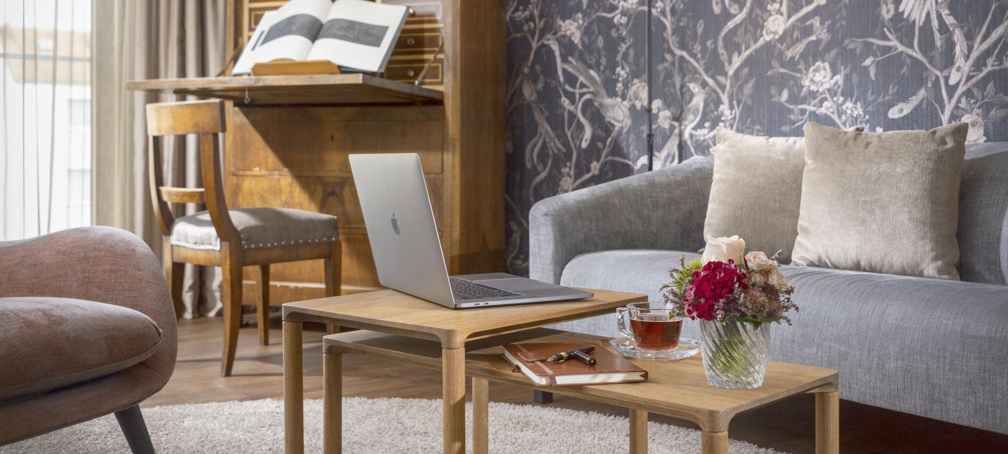 Wohnzimmer mit Laptop, Tee, Notizbuch und Blumen auf Holztisch; im Hintergrund Schreibtisch mit Buch – verbindet Arbeit und Gemütlichkeit.