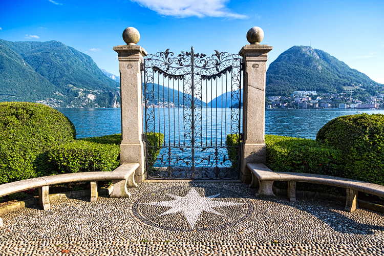 Blick auf den Luganersee mit Garten, Steinb&auml;nken und gr&uuml;nen Bergen im Hintergrund.