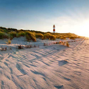  Sand dunes with grass and red-white lighthouse in sunlight under blue sky.