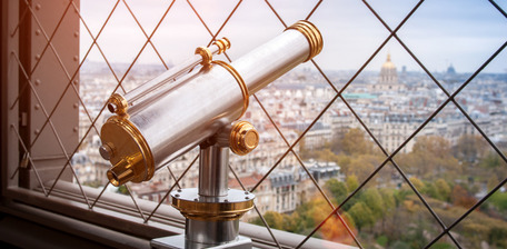 Aussichtsfernrohr mit Goldakzenten vor Stadtpanorama und Gitterzaun. 