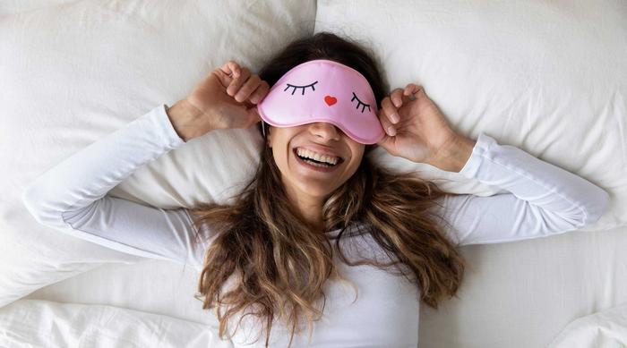  Person lies relaxed in bed, wearing a white shirt and holding a pink sleep mask. White background, calm sleeping atmosphere.