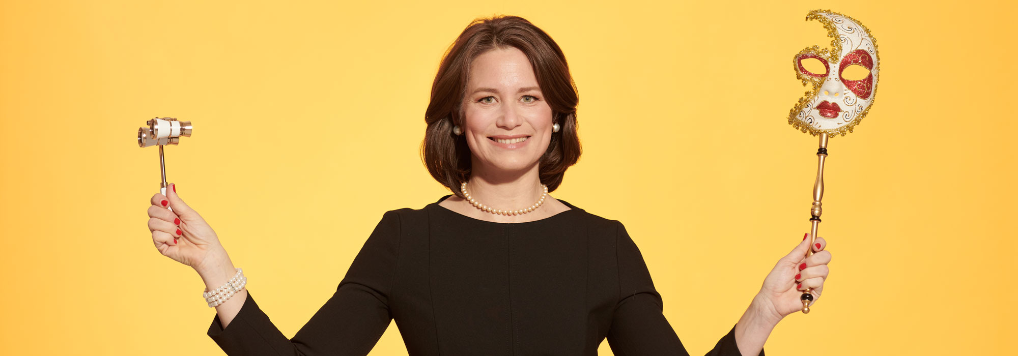 Sonja Wimmer hält Gavel und dekorative Maske vor gelbem Hintergrund.