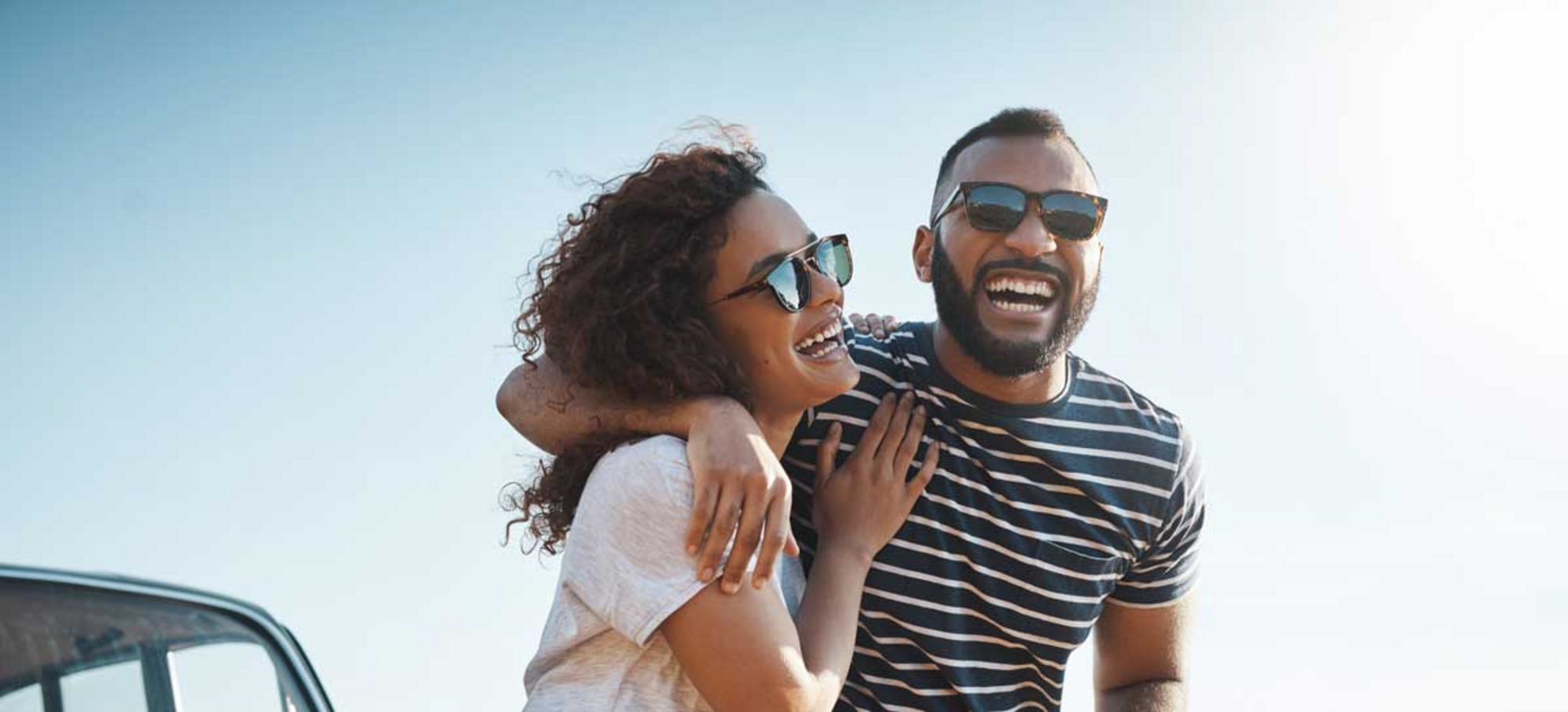 : Two people wearing sunglasses smile at the camera on a sunny day. 
