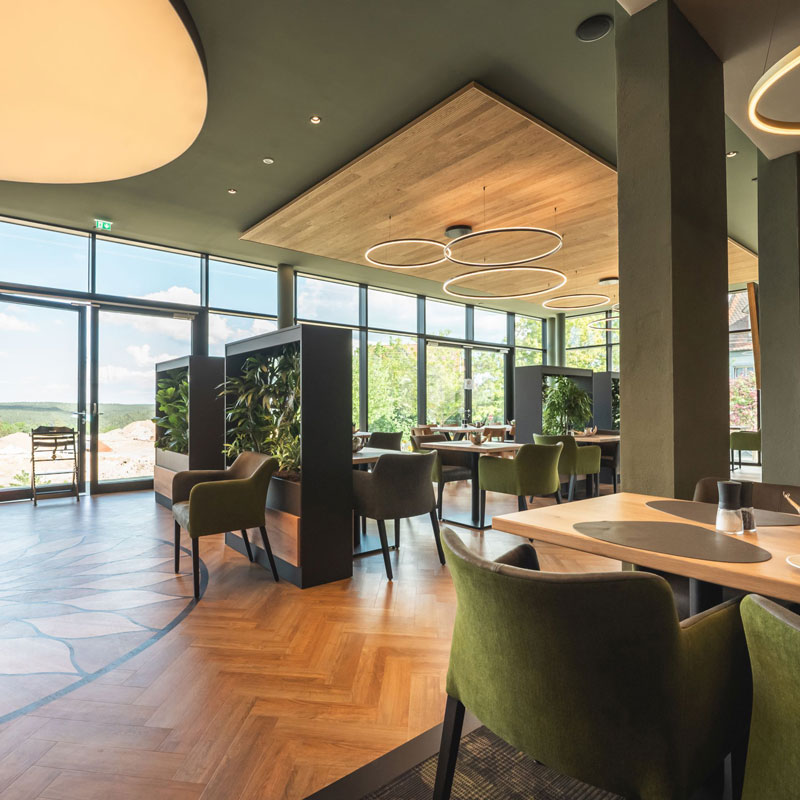 Modernes Restaurant mit gro&szlig;en Fenstern, gr&uuml;nen St&uuml;hlen und stilvollen Lampen.