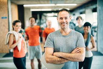 Group in gym, person in front smiling, others in background wearing workout clothes.