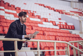 Person in suit with tablet in front of red-white stadium seats, leaning on railing.
