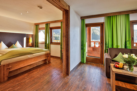 Gemütliches Hotelzimmer mit Bett, Sofa, Holzmöbeln, Blumen, Balkonblick auf Natur und grüne Vorhänge.