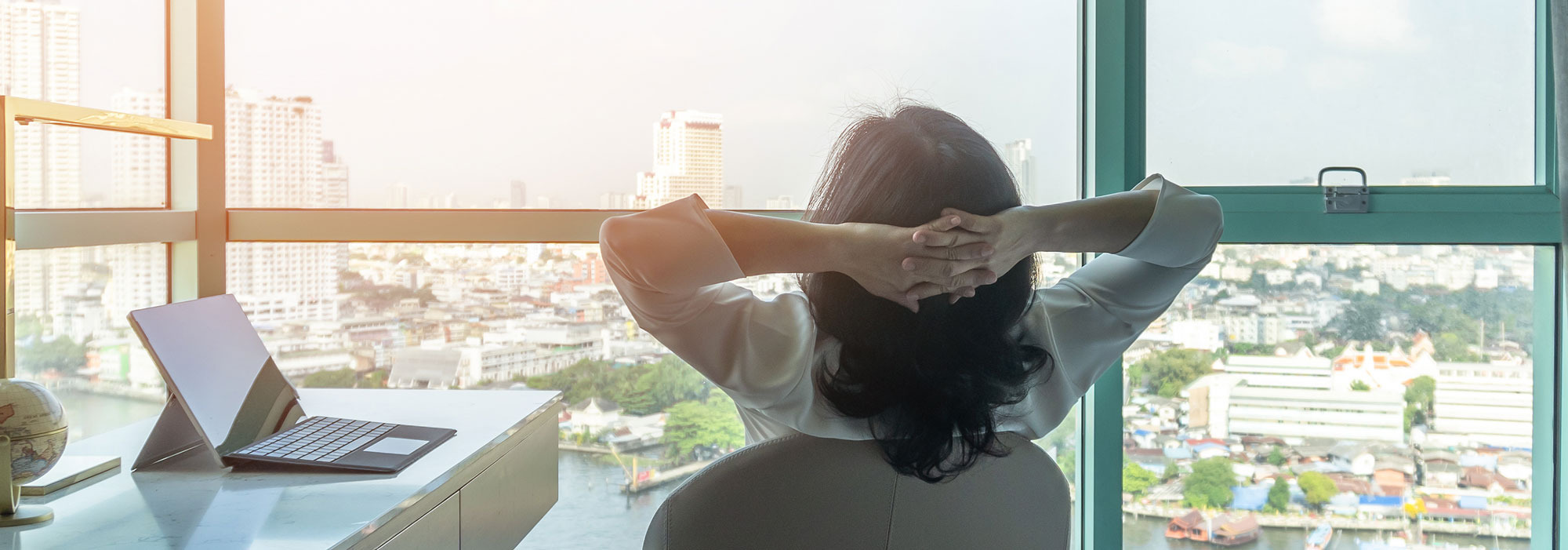 Person blickt aus Fenster auf Stadt, mit Laptop und Globus am Tisch – Symbol für Planung einer Geschäftsreise.