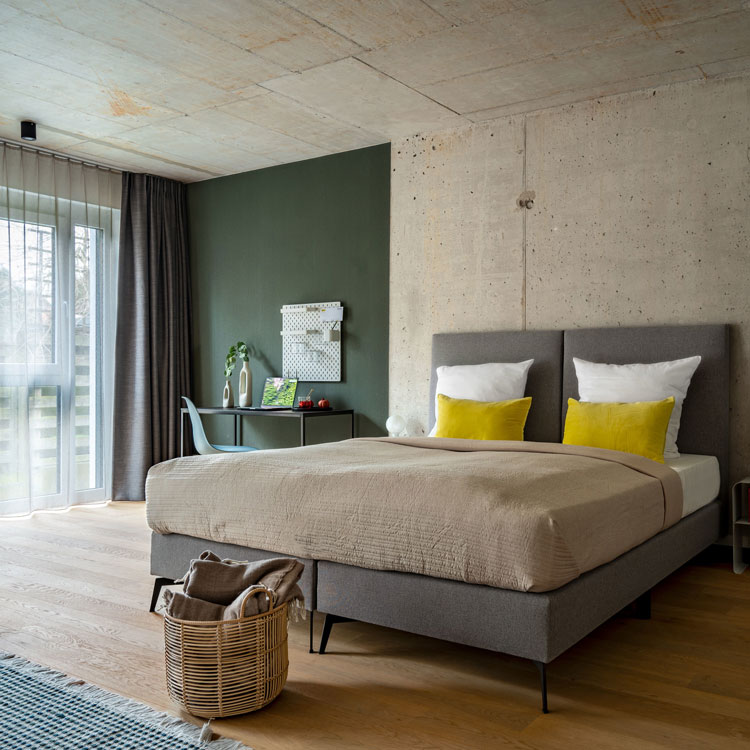 Modernes Schlafzimmer mit Doppelbett, Betonwand, Schreibtisch und gro&szlig;en Fenstern.