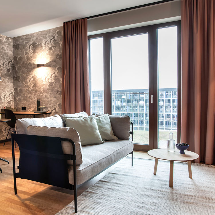 Modernes Wohnzimmer mit Sofa, Couchtisch, gro&szlig;en Fenstern und Schreibtisch im Hintergrund.