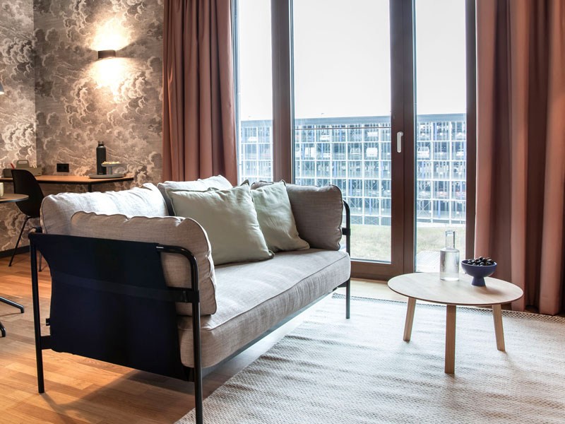 Modernes Wohnzimmer mit Sofa, Couchtisch, gro&szlig;en Fenstern und Schreibtisch im Hintergrund.