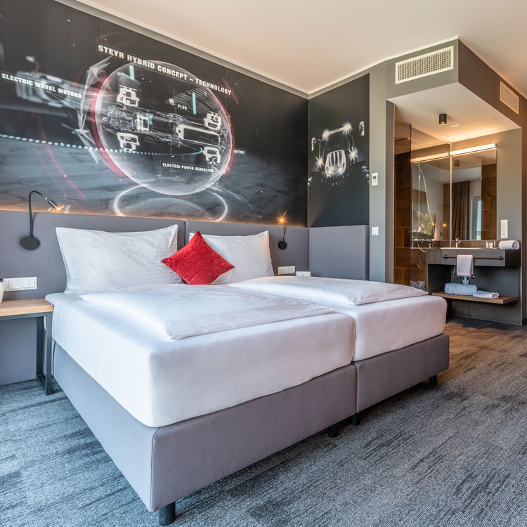 Modernes Hotelzimmer mit Doppelbett, rotem Zierkissen und Technik-Wandgrafik.