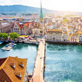 Zürich mit Limmat, historischen Gebäuden, grünen Hügeln und Sonnenstrahlen.
