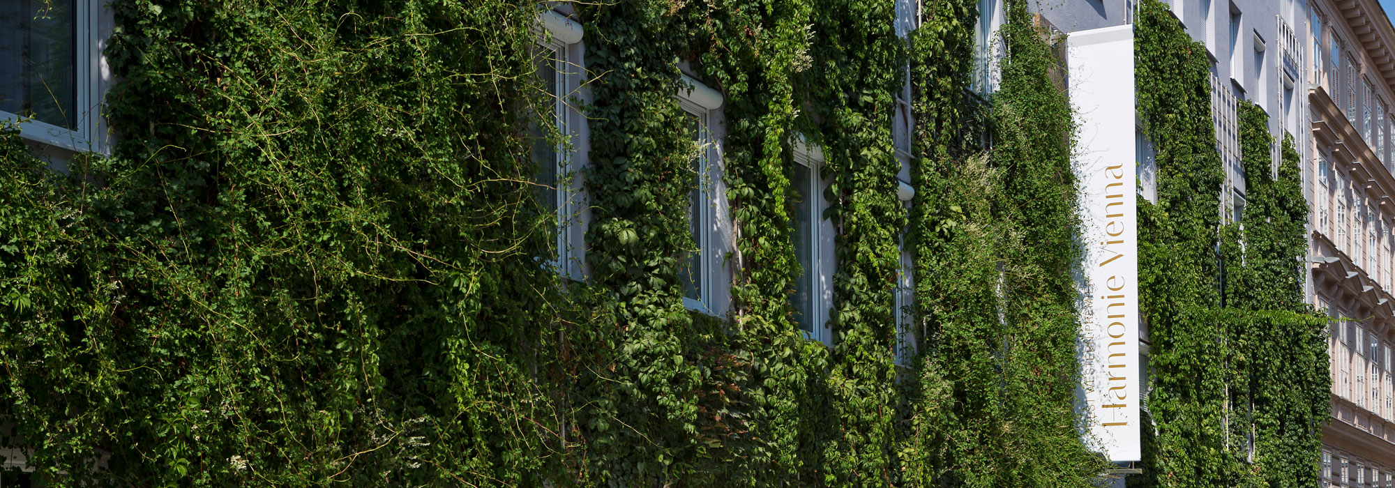 Geb&auml;udefassade dicht bewachsen mit gr&uuml;nem Pflanzenkleid und Schild &bdquo;Harmonie Vienna&ldquo;.