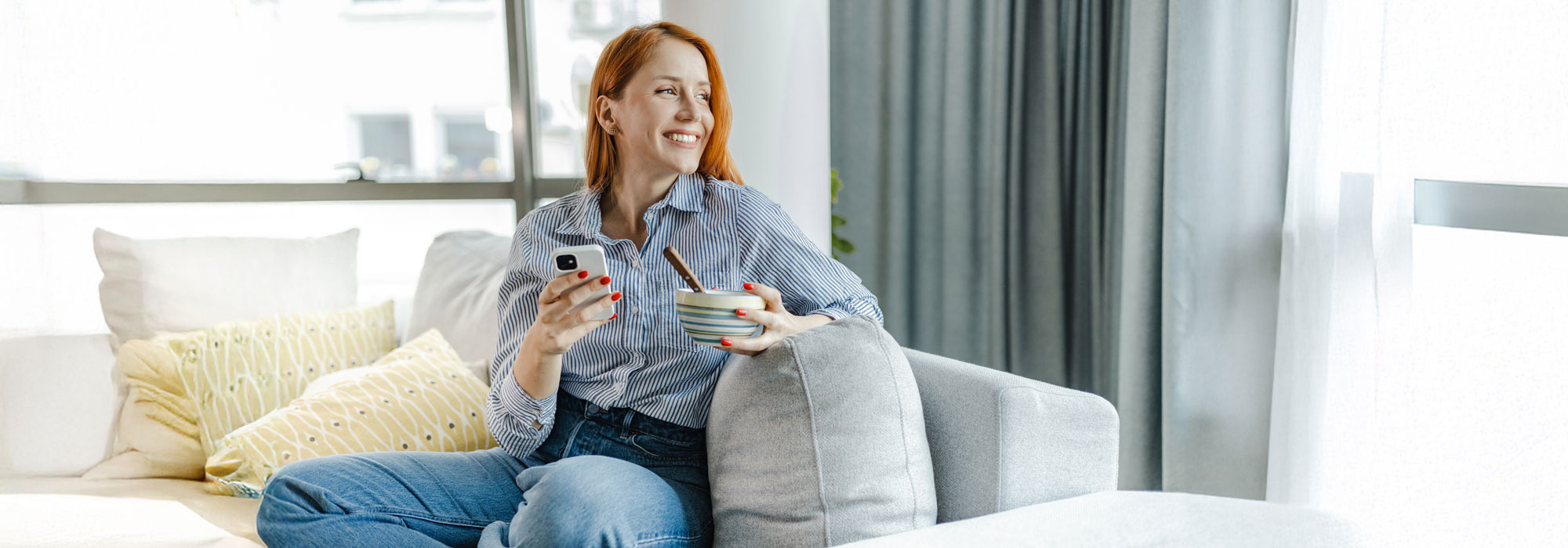 Person sitzt auf Sofa, hält Smartphone und Schüssel, helle moderne Wohnzimmereinrichtung.