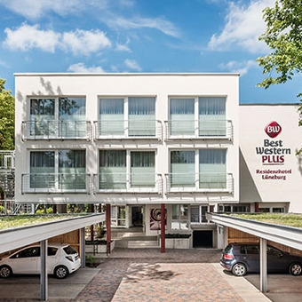 Außenansicht des Best Western Plus Residenzhotel Lüneburg mit weißer Fassade, Balkonen, Bäumen und überdachtem Parkplatz mit geparkten Autos.