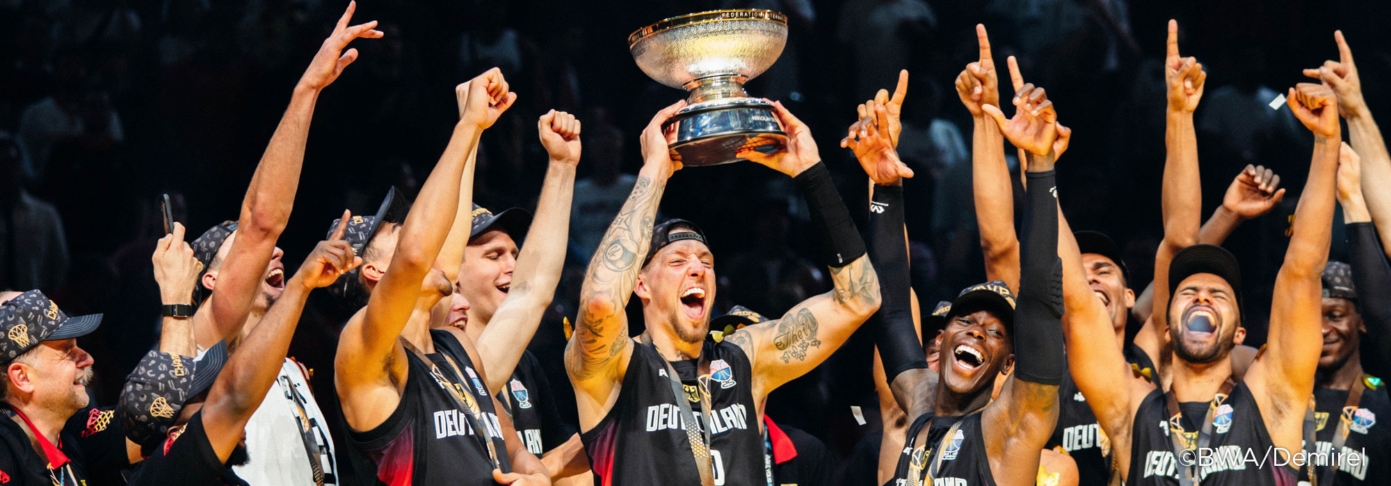 Basketball team celebrates with raised arms, holding a large trophy aloft.