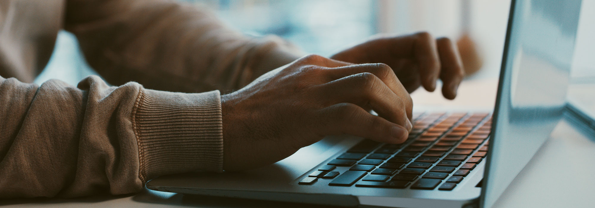 Zwei Hände tippen auf Laptop-Tastatur, unscharfer Hintergrund bei Tageslicht.