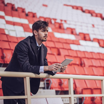 Person im Anzug mit Tablet vor rot-weißen Stadionplätzen, lehnt am Geländer.