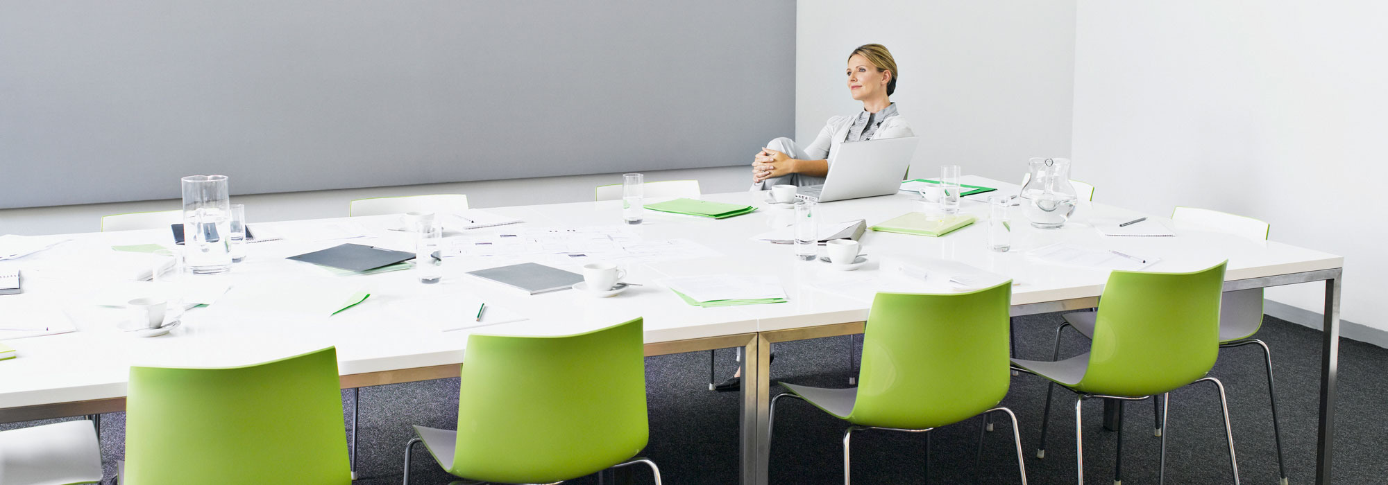 Person in modernem Konferenzraum mit weißem Tisch, grünen Stühlen und Laptop.
