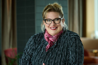 Carmen Dücker steht an einem Holztisch, trägt ein gestreiftes Oberteil und rosa Halstuch in modernem Raum.