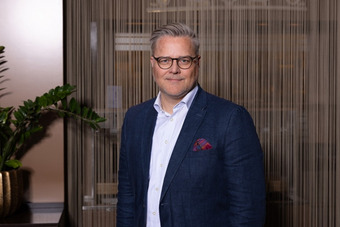 Carsten Weber standing in front of a partition, wearing a dark blue jacket and white shirt with pocket square.