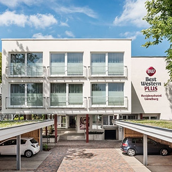 Exterior view of the Best Western Plus Residenzhotel Lüneburg with white facade, balconies, trees, and covered parking area with parked cars.