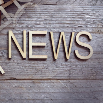 Große Holzbuchstaben formen das Wort ‚NEWS‘ auf grauem Holzhintergrund, weitere Buchstaben liegen gestapelt oben links im Bild