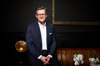 Marcus Smola seated on a brown leather couch, wearing a dark suit against a black wall background.
