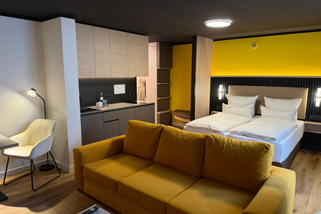 Modernes Apartment mit gelbem Sofa, Küchenzeile und Doppelbett vor gelber Wand.